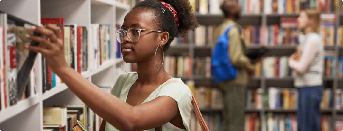 Lady wearing reading glasses picking a book from the library shelf to gain some financial knowledge.